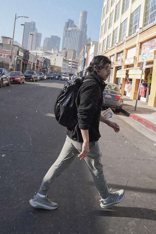 Dr. Rab, wearing glasses, a black fleece jacket, and grey pants, walks across a downtown street while carrying a black backpack, with the Los Angeles skyline visible in the background.