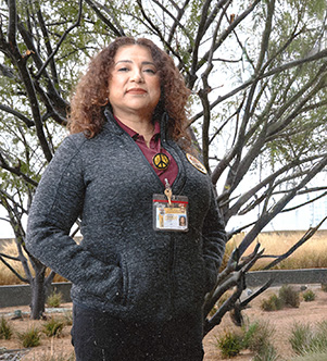 Amparo stands outdoors with hands in her pockets, wearing a grey zip-up jacket with a peace-sign pin over a maroon collared shirt and a County of Los Angeles ID lanyard, with bare branching trees and dry grasses in the background.