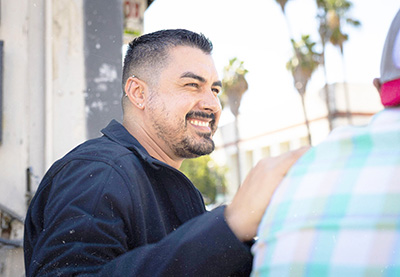 Carlos smiles warmly while in conversation with a client whose back is to the camera, wearing a black zip-up jacket, with palm trees and a sunny sky in the background.
