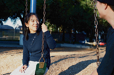 Dr. Yamaguchi sits on a swing at a playground, smiling warmly while in conversation with another person whose back is to the camera, wearing a dark knit sweater, with trees and woodchip ground visible in the background.