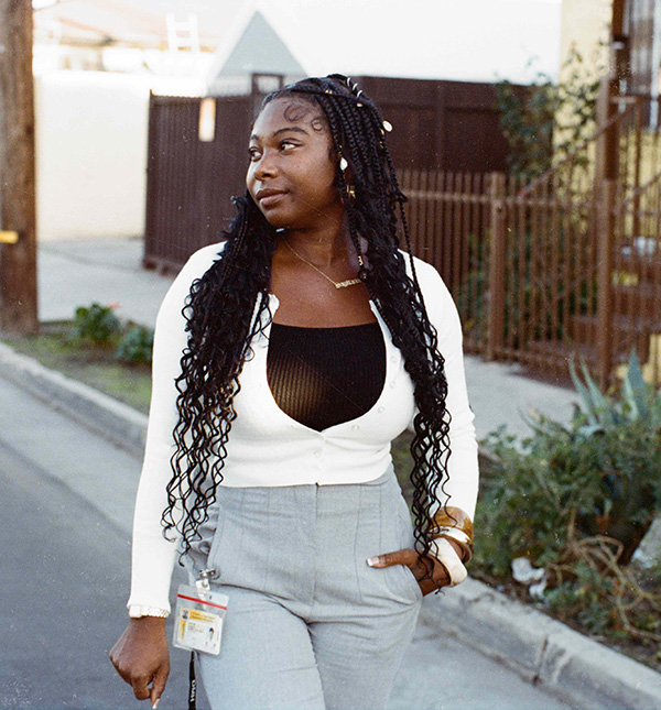 Cyndi stands outdoors looking off to the side, wearing a cream button-up cardigan over a black top, grey trousers, a gold name necklace, and a County ID lanyard, with a residential gate and greenery in the background.