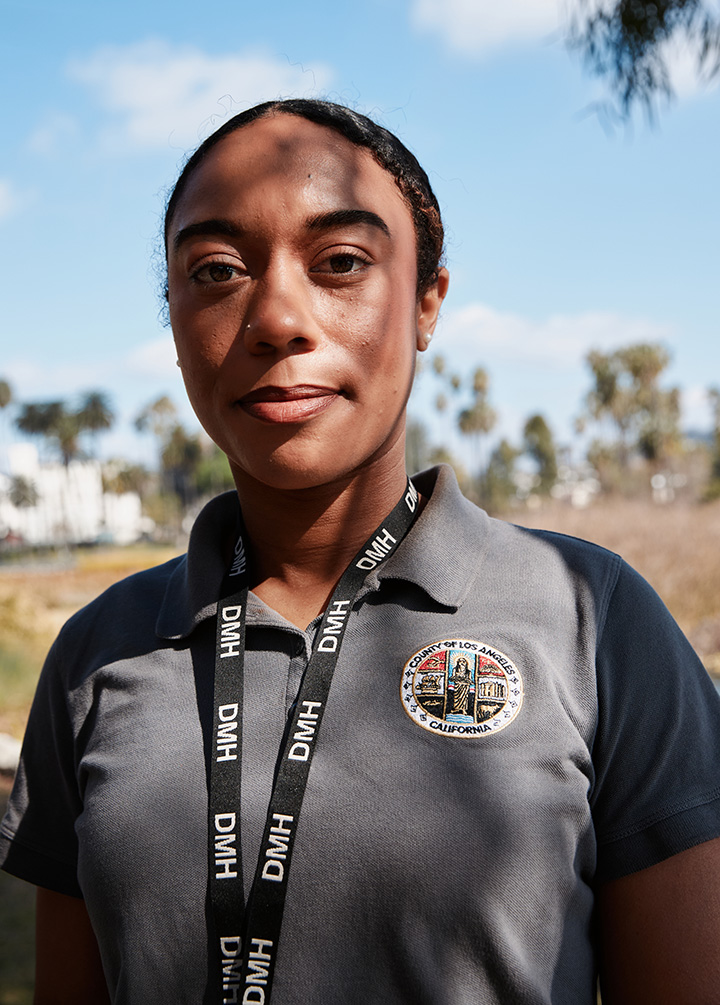 Aleetia looks directly at the camera with a calm expression, wearing a grey polo shirt with a County of Los Angeles patch and a black DMH lanyard, with palm trees and a blue sky in the background.
