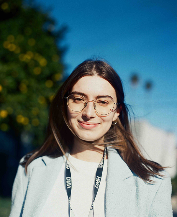 Albina smiles softly at the camera in a close-up portrait, wearing round gold-framed glasses, a light blue blazer over a white top, and a black DMH lanyard, with a lemon tree and palm trees visible in the sunny background.