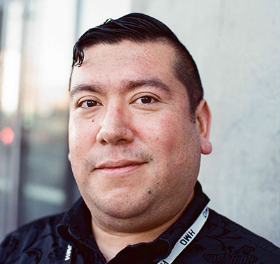 Daniel smiles softly at the camera in a close-up portrait, wearing a dark patterned button-up shirt and a black DMH lanyard, with a light grey wall in the background.