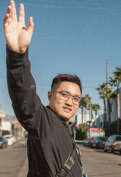 Josh smiles at the camera with his hand raised in a wave, wearing glasses, a black jacket, and a crossbody bag strap, with palm trees, parked cars, and a sunny street in the background.
