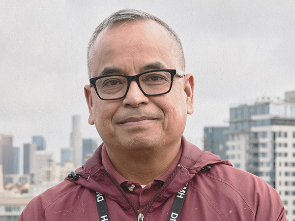 Leo smiles softly at the camera in a close-up portrait, wearing black-framed glasses, a maroon zip-up jacket, and a black DMH lanyard, with the Los Angeles skyline blurred in the background.