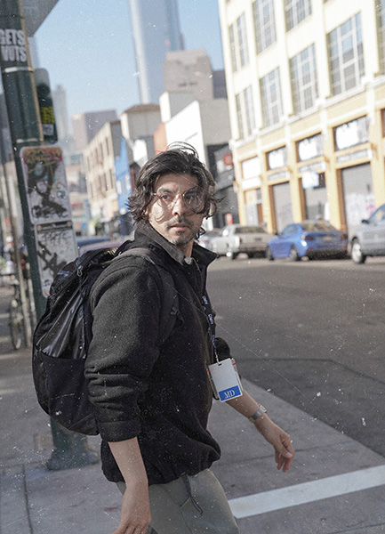 Dr. Rab glances over his shoulder toward the camera at a sunlit downtown intersection, wearing round glasses, a black jacket, a DMH lanyard, and a black backpack, with storefronts, traffic signals, and a 4th Street sign visible in the background.