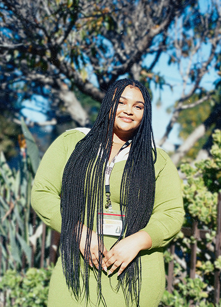 Zariah smiles softly at the camera, wearing an olive-green sweater over a white collared shirt with a DMH ID lanyard, her long braided hair flowing down, with trees and greenery in the sunlit background.