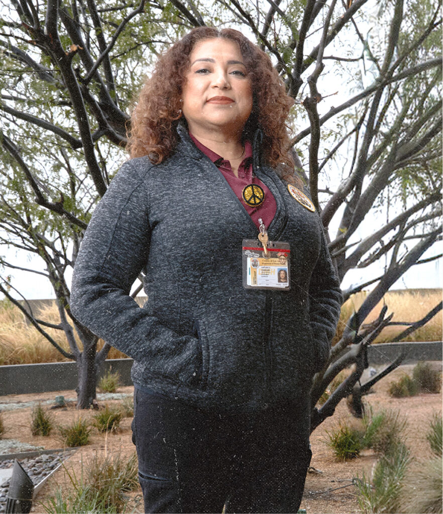 Amparo stands outdoors with hands in her pockets, wearing a grey zip-up jacket with a peace-sign pin over a maroon collared shirt and a County of Los Angeles ID lanyard, with bare branching trees and dry grasses in the background.