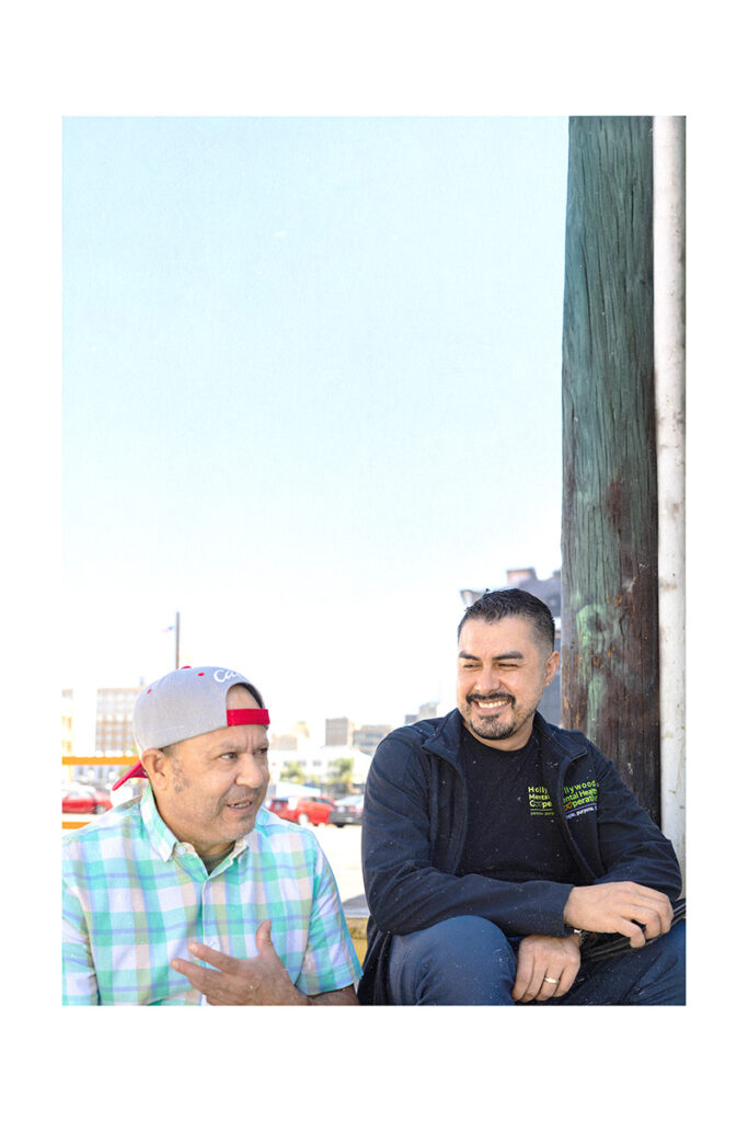 Carlos sits outdoors smiling as he listens to a client in conversation, wearing a black Hollywood Mental Health Cooperative zip-up jacket, with a wooden utility pole and a sunny urban backdrop behind them.