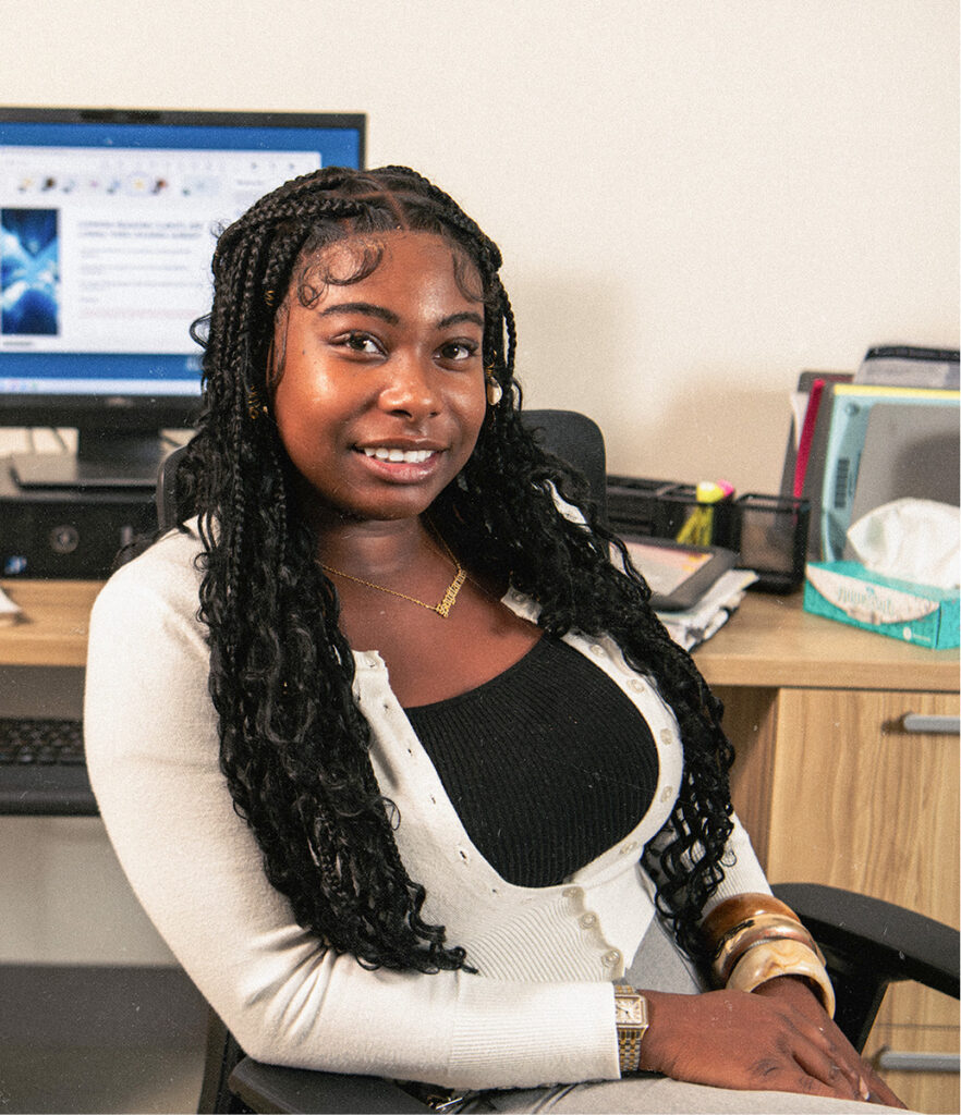 Cyndi smiles at the camera while seated at an office desk, wearing a cream button-up cardigan over a black top with a gold name necklace and watch, with a computer monitor and office supplies visible behind her.