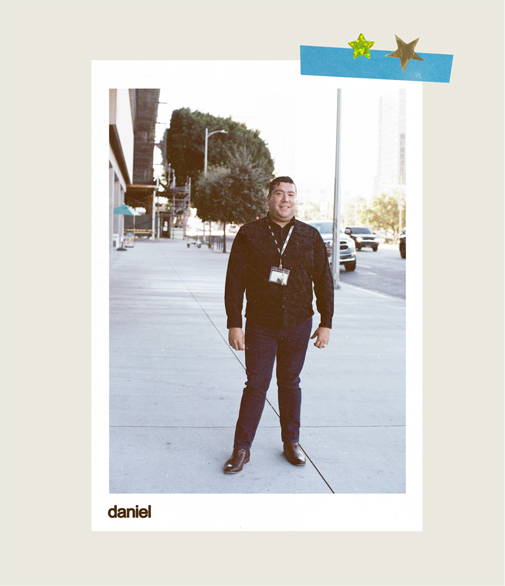 Daniel stands on a city sidewalk smiling at the camera, wearing a dark button-up shirt with an ID lanyard, dark pants, and brown leather boots, presented as a Polaroid-style photo labelled "Daniel" with decorative star stickers in the corner.