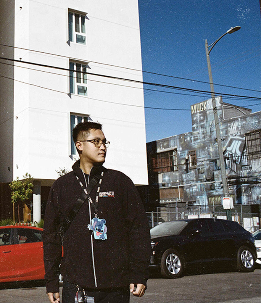 Josh stands outdoors looking off to the side, wearing glasses, a black jacket, a crossbody bag strap, and a DMH lanyard with a Stitch-character ID holder, with a tall white building, a mural, and parked cars visible in the background.