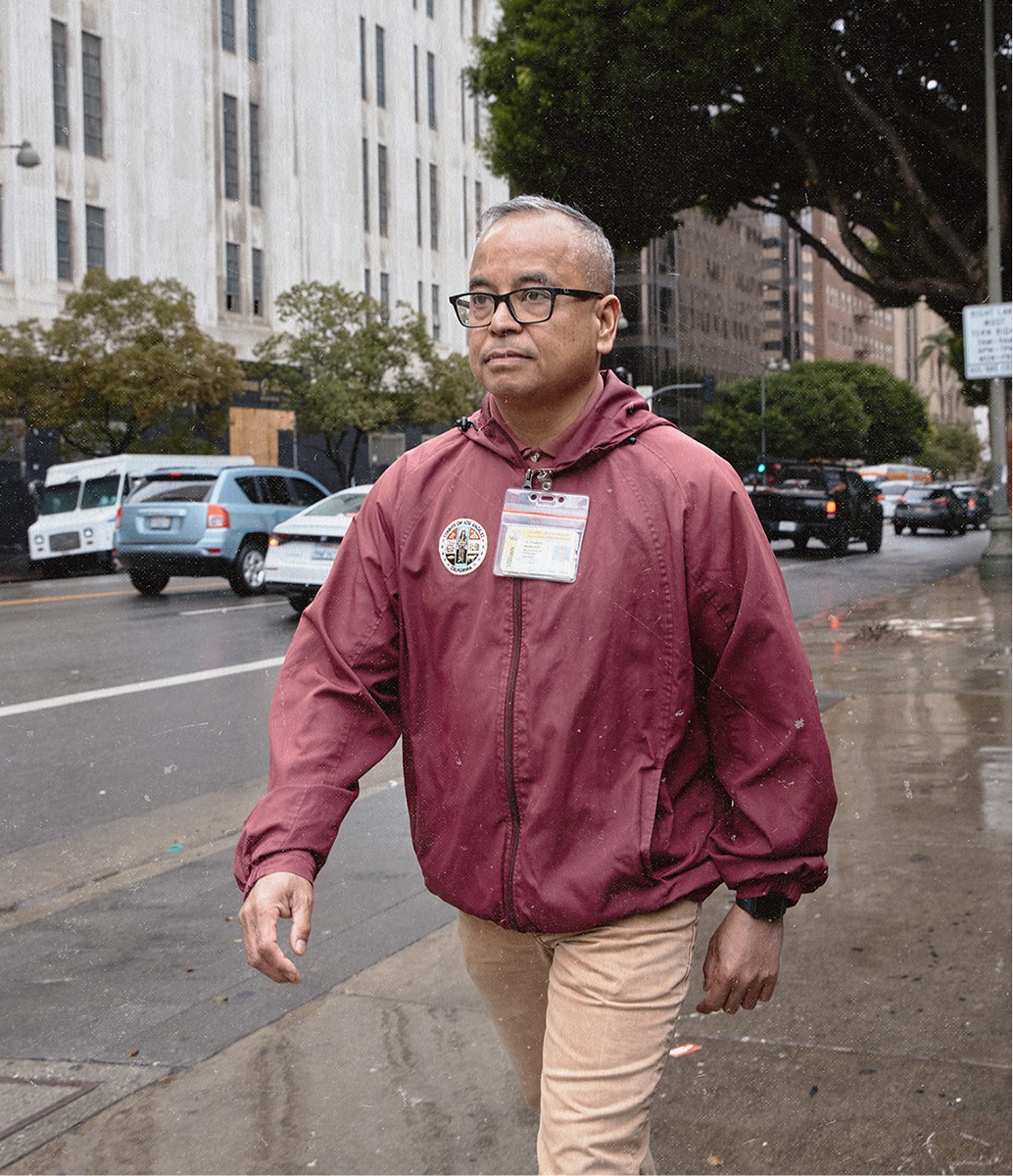 Leo walks along a wet city sidewalk wearing glasses, a maroon zip-up jacket with a County of Los Angeles patch and ID lanyard, and khaki pants, with traffic and a large municipal building visible in the background.