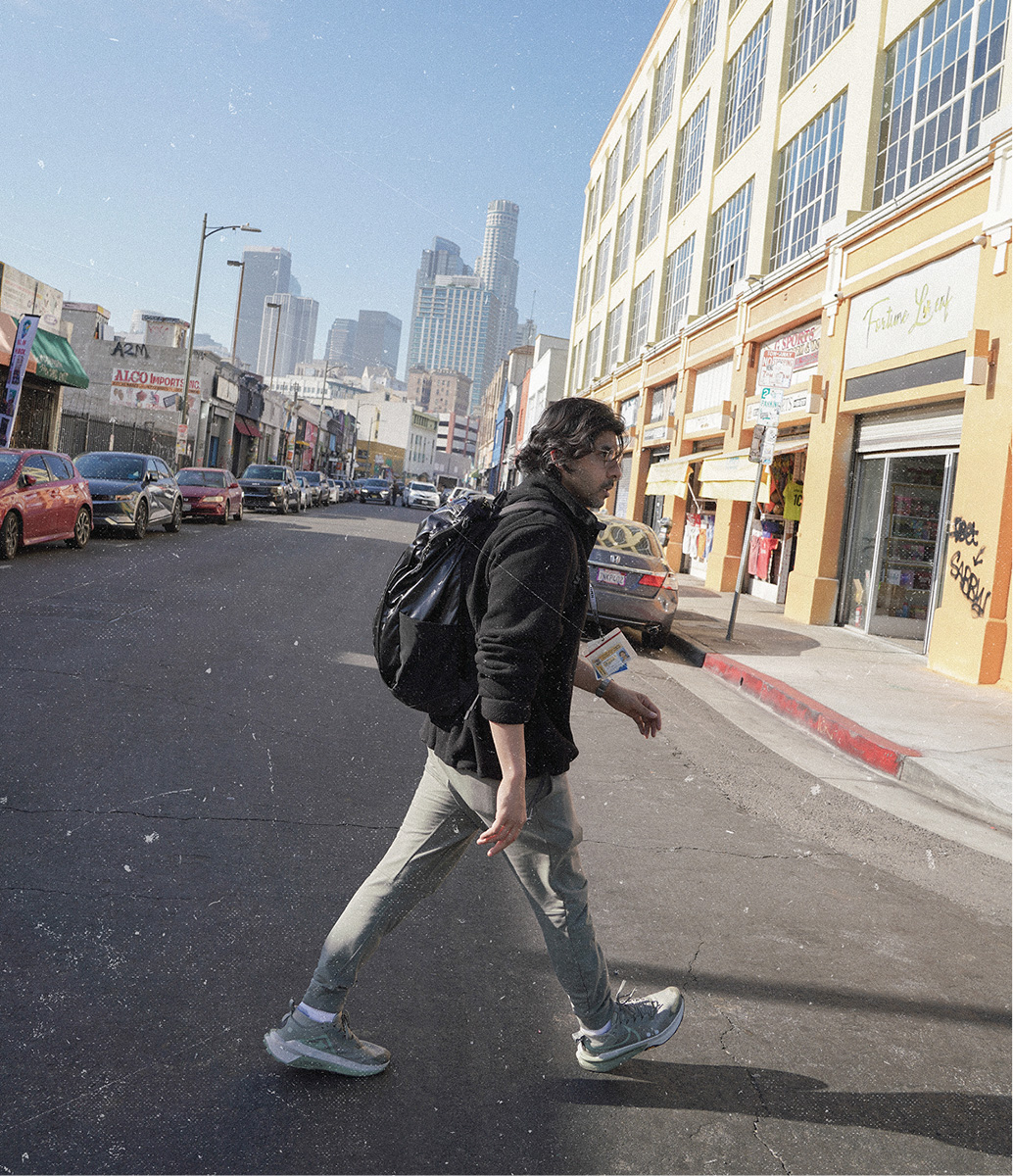 Dr. Rab, wearing glasses, a black fleece jacket, and grey pants, walks across a downtown street while carrying a black backpack, with the Los Angeles skyline visible in the background.