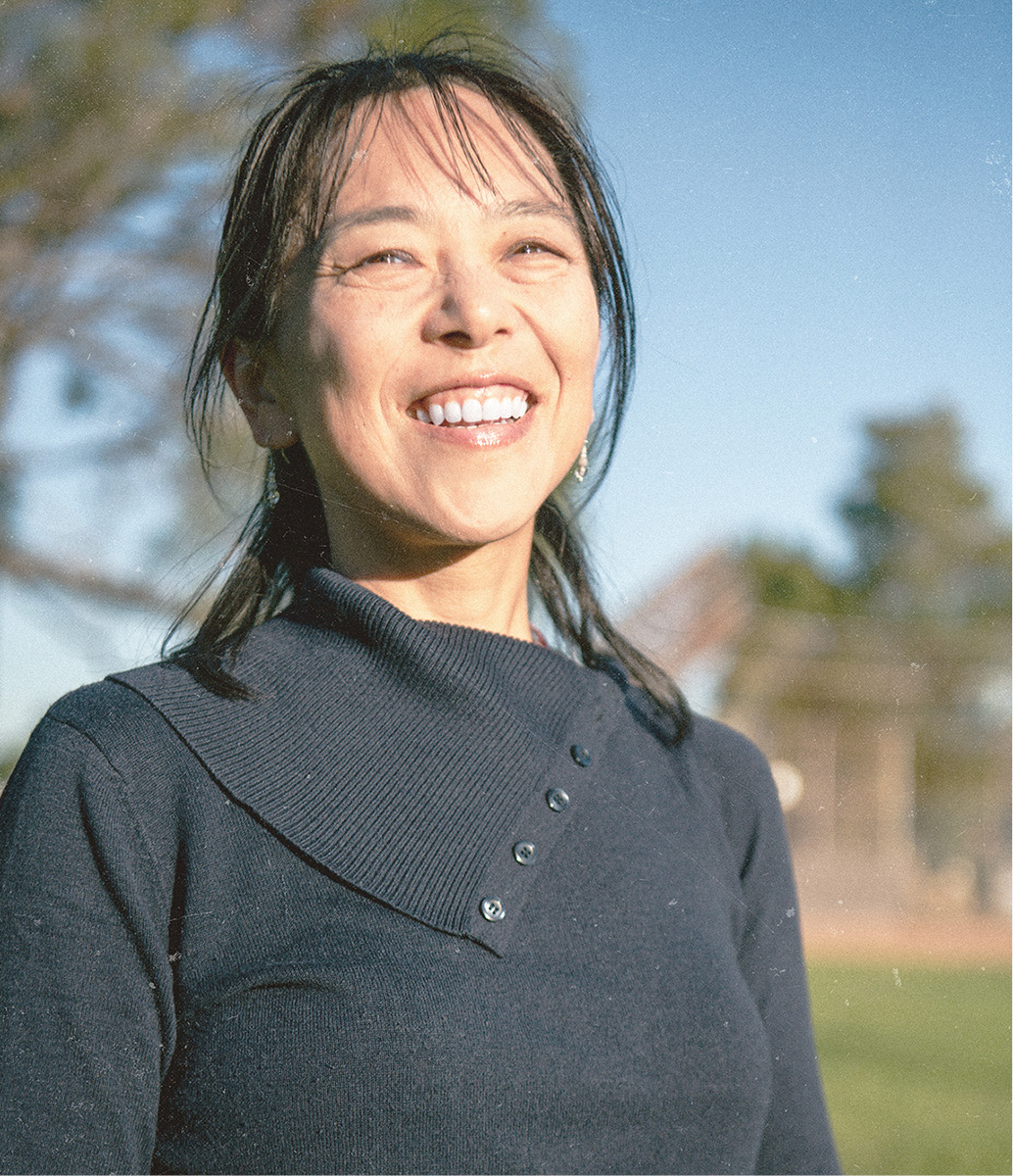 Dr. Yamaguchi smiles brightly while looking off to the side, wearing a dark knit sweater with an asymmetrical buttoned collar, photographed outdoors with trees and blue sky in the soft background.