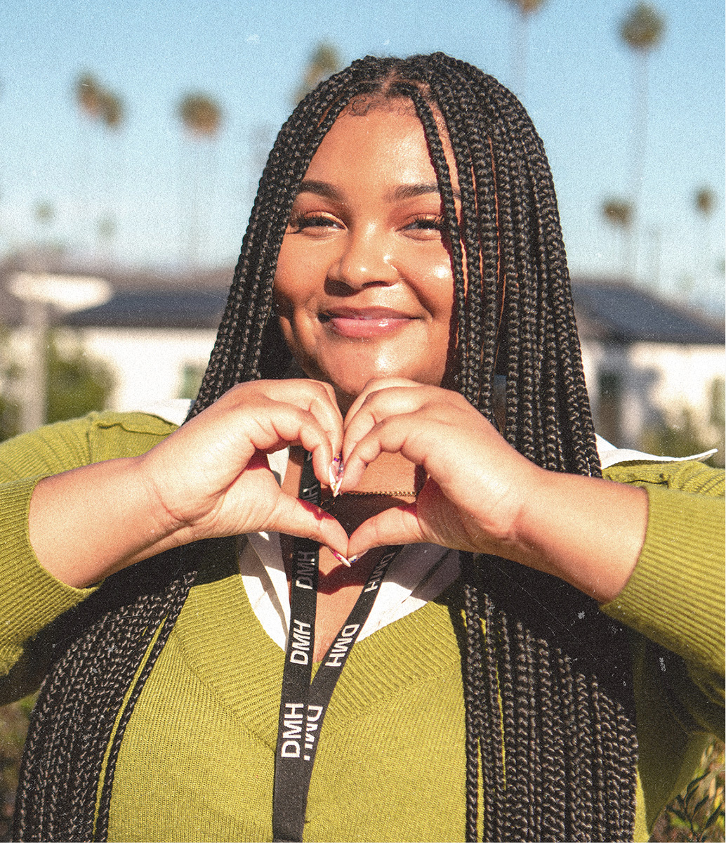 Zariah smiles at the camera while forming a heart shape with her hands, wearing an olive-green sweater over a white collared shirt and a black DMH lanyard, with long braided hair and palm trees visible in the sunny background.